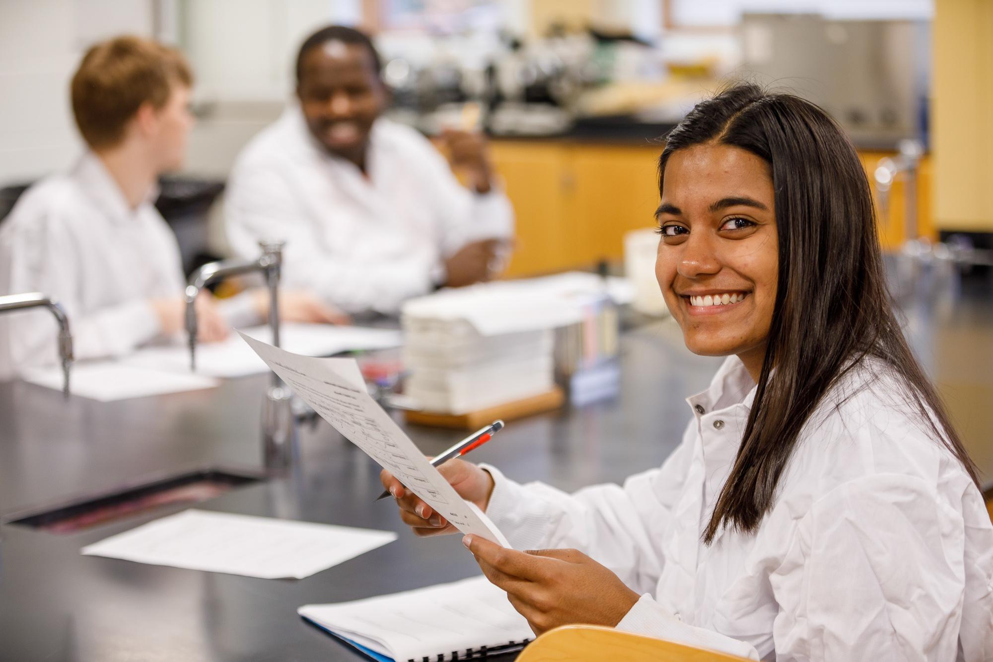 Woman in a lab coat smiling while holding a paper, two students talk behind her